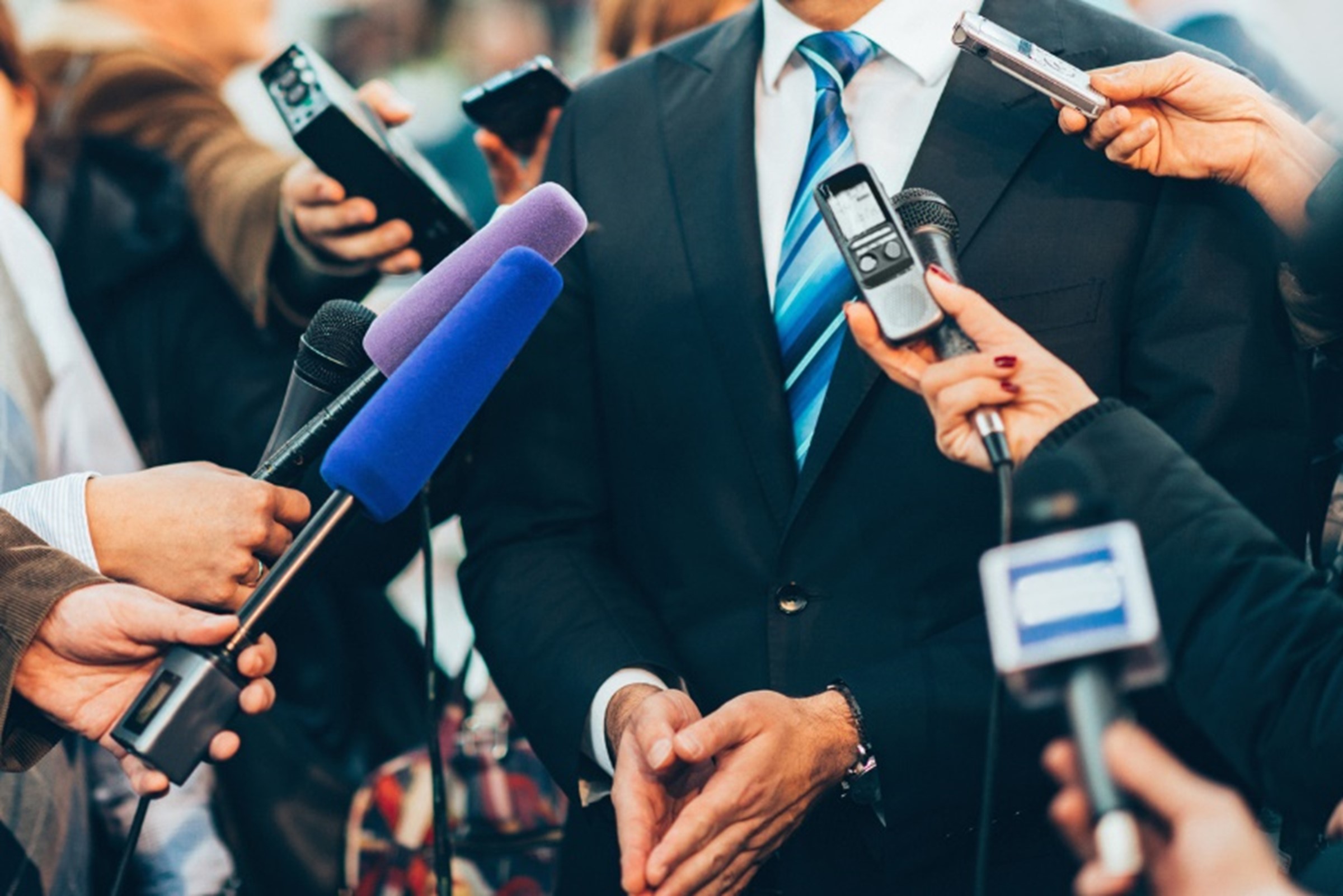 a man in a suit from neck down with microphones and people around him